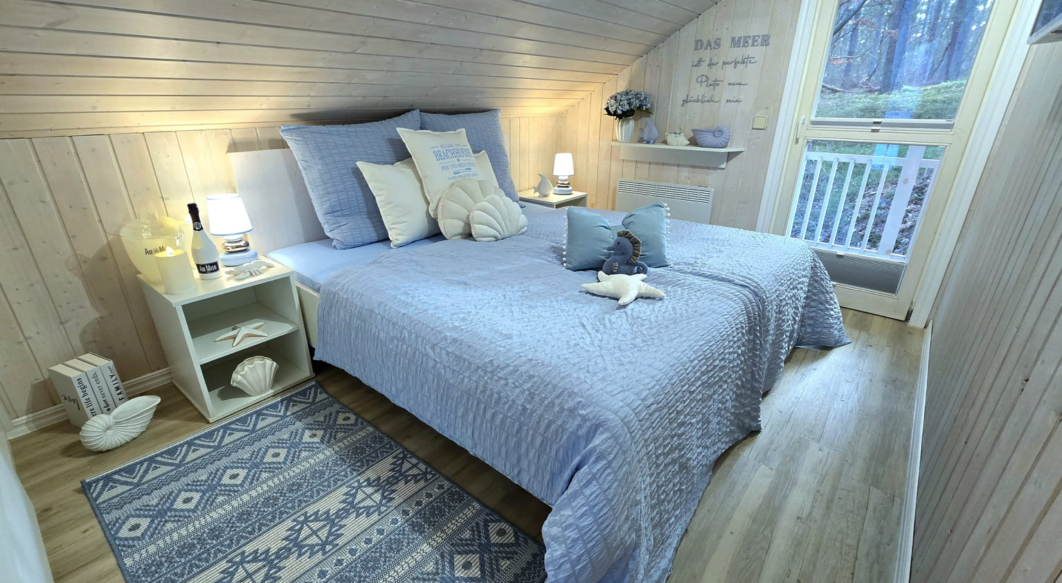 Gemütliches Schlafzimmer mit Dachschräge, einem Doppelbett mit blauer Bettwäsche, einem Nachttisch mit Lampe und Dekorationen, einem Teppich und einem Fenster mit Blick nach draußen.